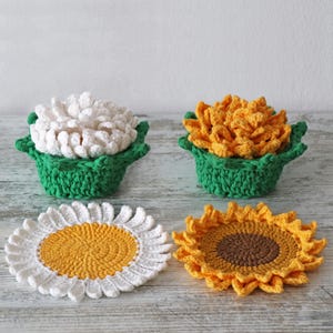 Op de afbeelding: Vier handgemaakte gehaakte onderzetters in een bloemig thema. Twee onderzetters zijn gevormd als madeliefjes, met witte bloemblaadjes en gele centra. De andere twee zijn 3D bloemontwerpen, één wit en één geel, in groene gehaakte manden.