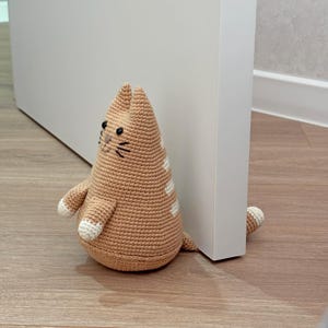 May include: A light brown crocheted cat doorstop with white accents on the paws and back. The cat features black embroidered eyes and whiskers, a triangular body shape, and is positioned to hold a white door open on a wooden floor surface.