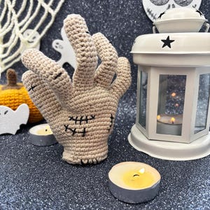May include: A beige crochet hand with black stitched details, resembling a zombie hand, is the focal point. The hand is surrounded by Halloween decorations, including a white lantern, candles, and a small pumpkin. The background is a glittery dark surface.