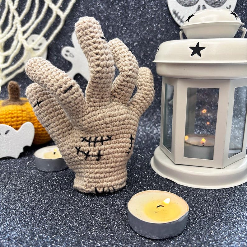 May include: A beige crochet hand with black stitched details, resembling a zombie hand, is the focal point. The hand is surrounded by Halloween decorations, including a white lantern, candles, and a small pumpkin. The background is a glittery dark surface.