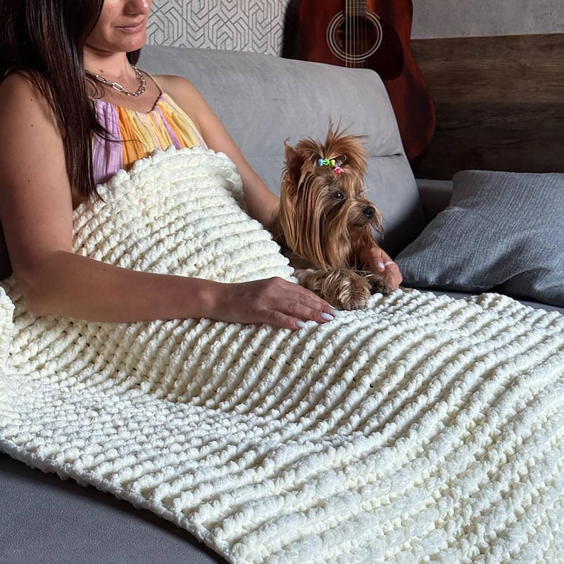 May include: A cream-colored, chunky knit blanket with a textured, ridged pattern. The blanket is draped over a gray couch, with a small brown dog with a topknot resting on top. The dog has a colorful hair accessory. A guitar is visible in the background.