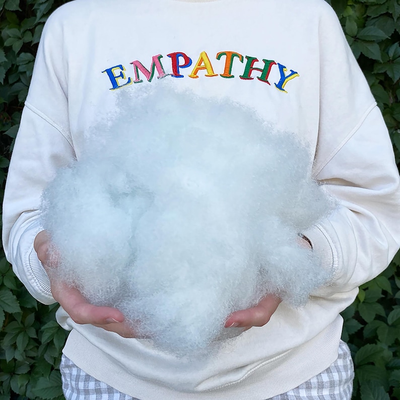 May include: A person wearing a cream-colored sweatshirt with the word "EMPATHY" embroidered in colorful letters, holding a large cloud of fluffy, white stuffing. The background is a green leafy plant.