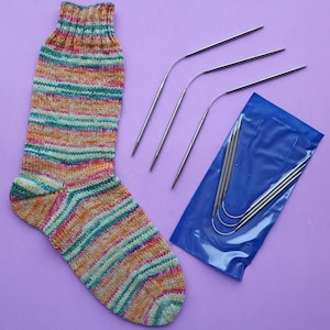 Könnte beinhalten: Eine bunte gestreifte Socke in Grün-, Orange- und Rosatönen. Die Socke liegt auf einer violetten Oberfläche neben einer Packung mit vier silbernen Stricknadeln.