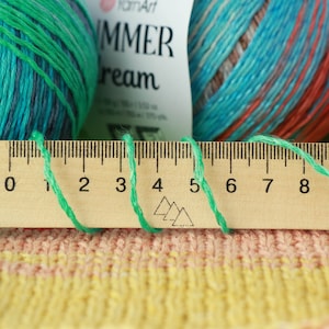 May include: A wooden ruler with black markings and numbers from 0 to 8. Green yarn is wrapped around the ruler, creating a loop shape.