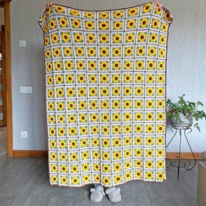 K&ouml;nnte beinhalten: Eine gro&szlig;e, handgefertigte H&auml;keldecke mit einem sich wiederholenden Sonnenblumenmuster. Jedes Quadrat zeigt eine gelbe Sonnenblume mit brauner Mitte und gr&uuml;nen Bl&auml;ttern auf wei&szlig;em Hintergrund. Die Decke hat einen braunen Rand.