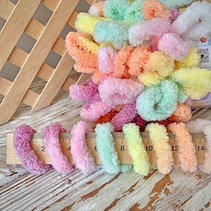 May include: A collection of colorful, fluffy yarn in pastel shades of pink, purple, yellow, green, and orange. The yarn is measured with a wooden ruler, showing lengths up to 14 inches. The yarn is arranged near a wooden lattice.