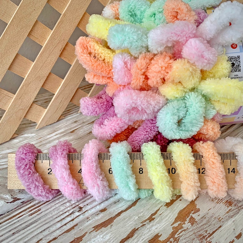 May include: A collection of colorful, fluffy yarn in pastel shades of pink, purple, yellow, green, and orange. The yarn is measured with a wooden ruler, showing lengths up to 14 inches. The yarn is arranged near a wooden lattice.