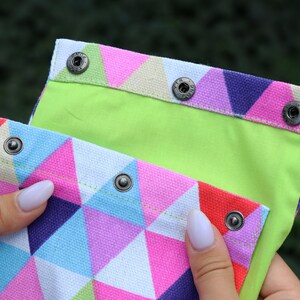 May include: A colorful fabric pouch with a geometric pattern of pink, blue, and white triangles. The pouch has a lime green lining and a flap closure with silver snaps.