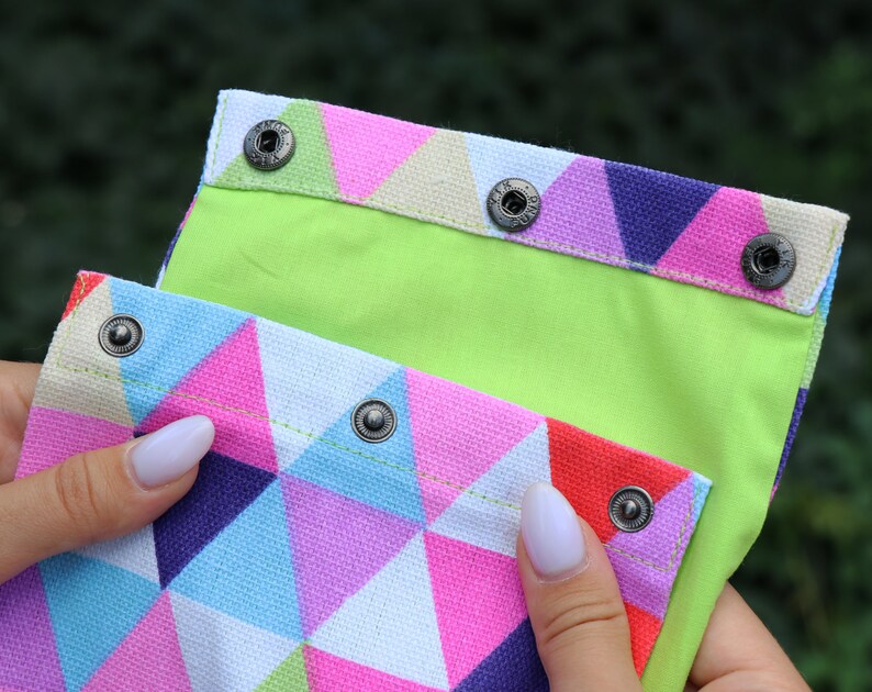 May include: A colorful fabric pouch with a geometric pattern of pink, blue, and white triangles. The pouch has a lime green lining and a flap closure with silver snaps.