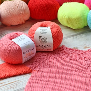 May include: A collection of colorful yarn balls in various shades, including pink, red, and blue, with a partially knitted pink baby sweater. The yarn labels read "GAZZAL BABY COTTON" and indicate a blend of cotton and acrylic.