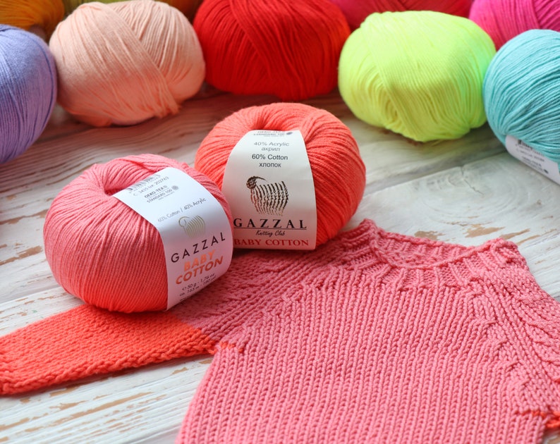May include: A collection of colorful yarn balls in various shades, including pink, red, and blue, with a partially knitted pink baby sweater. The yarn labels read "GAZZAL BABY COTTON" and indicate a blend of cotton and acrylic.