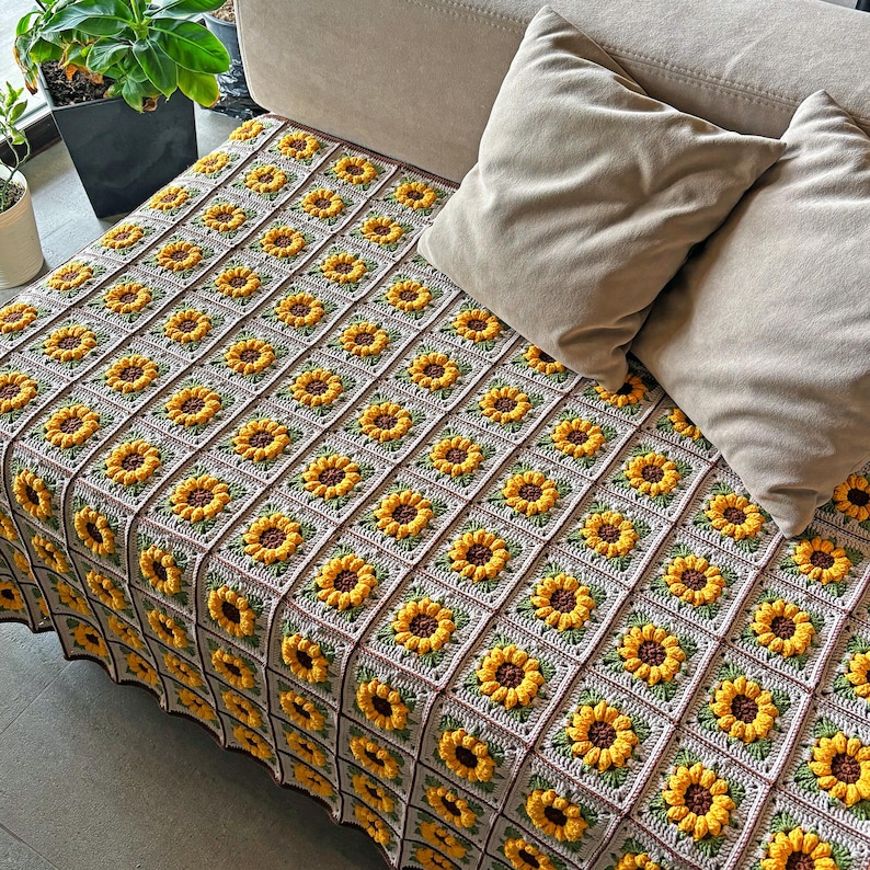 K&ouml;nnte beinhalten: Eine handgefertigte H&auml;keldecke mit einem sich wiederholenden Sonnenblumenmuster. Die Decke besteht aus Quadraten mit gelben Sonnenblumen, braunen Zentren und gr&uuml;nen Bl&auml;ttern. Der Hintergrund ist hellgrau.