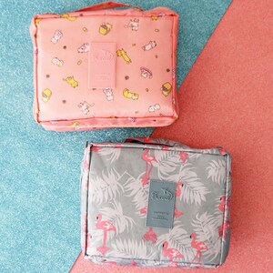 May include: Two travel cosmetic bags. One is pink with a cat and food print and the other is grey with a flamingo and leaf print. Both bags have a "Travel" logo.