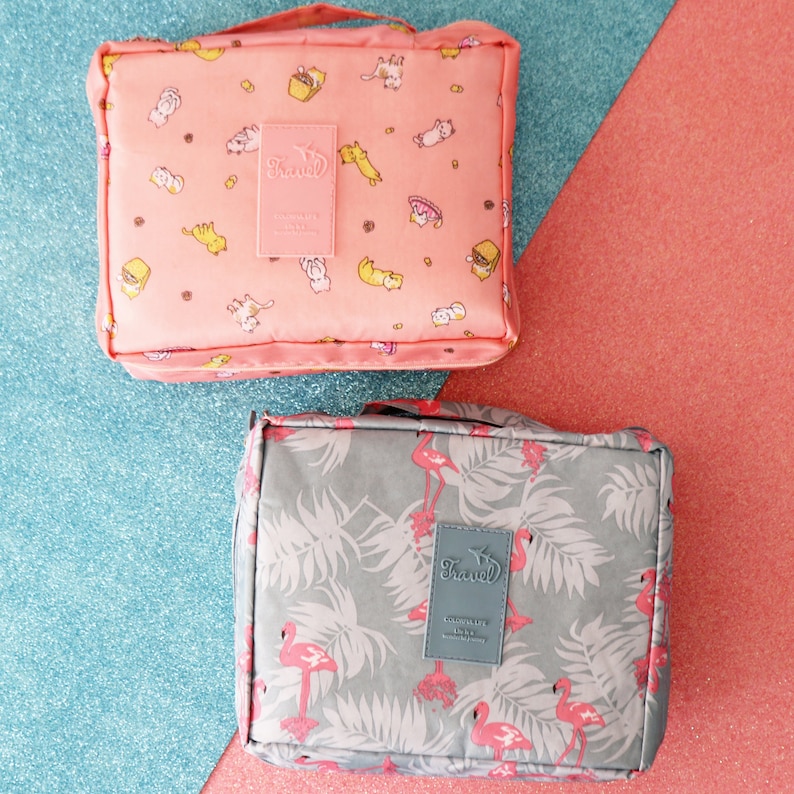 May include: Two travel cosmetic bags. One is pink with a cat and food print and the other is grey with a flamingo and leaf print. Both bags have a "Travel" logo.