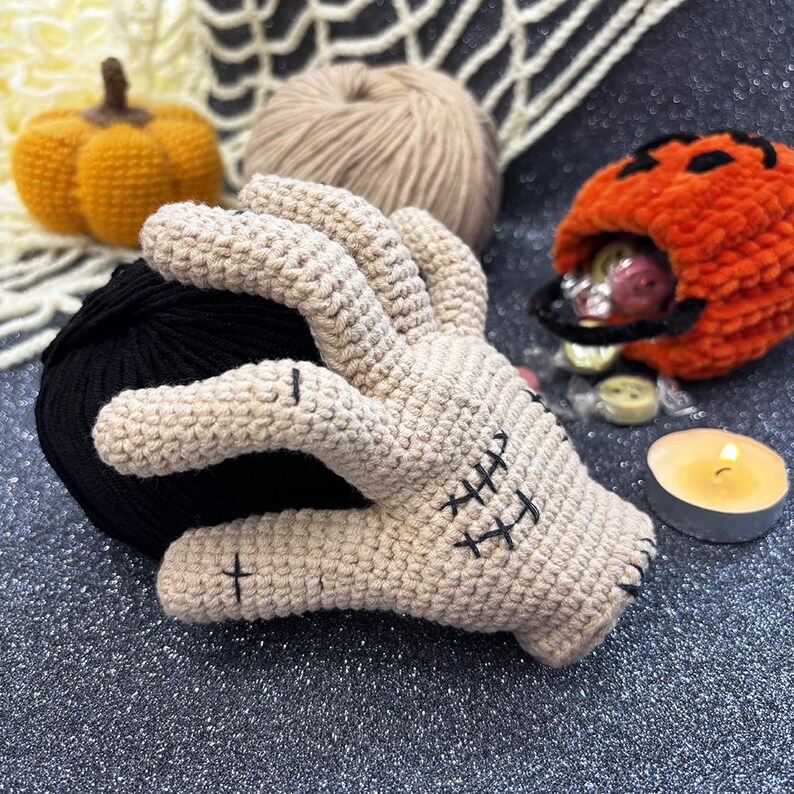 May include: A beige crochet hand with black stitched details, next to a black yarn ball, a small orange pumpkin, and a pumpkin-shaped candy holder. A lit candle and a ball of tan yarn are also in the image. Halloween-themed.