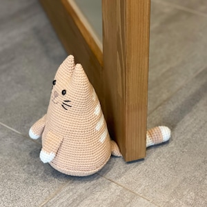 Puede incluir: Un tope de puerta de gato de ganchillo hecho a mano, de color melocotón. El gato tiene rayas blancas, ojos negros y bigotes. La cola es a rayas melocotón y blanco. El gato está colocado junto a un marco de puerta de madera.