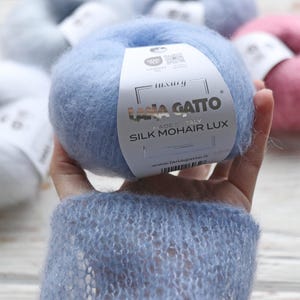 May include: A skein of light blue Lana Gatto Silk Mohair Lux yarn. The label reads "luxury" and "Lana Gatto" with a silver background. The label also says "Made in Italy" and "Silk Mohair Lux".