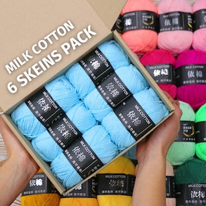 Op de afbeelding: Een doos met zes strengen lichtblauw Milk Cotton garen, met etiketten in Chinese karakters. De doos wordt door een persoon vastgehouden, met andere kleurrijke garenstrengen op de achtergrond, waaronder roze, groen en geel.