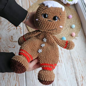 May include: A brown crocheted gingerbread man toy with white icing and red trim. The toy has black eyes and a white snowflake design on its chest.
