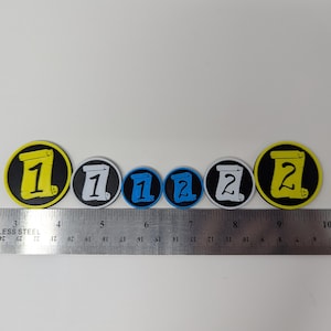May include: A set of six round game tokens with black, white, yellow, and blue colors. The tokens feature a scroll design with the numbers 1 and 2 printed on them.