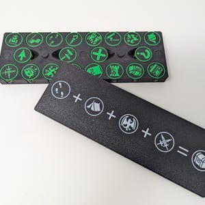 May include: A black plastic storage case with a lid, containing small green and white tokens with various symbols. The lid has a black and white graphic of a tent, footprints, a dragon, a sword, and a chest.