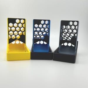 May include: Three colorful, geometric phone stands. Each stand has a rectangular base and a honeycomb-patterned back. The stands are yellow, blue, and black, with a black honeycomb design. The stands are designed to hold a phone.