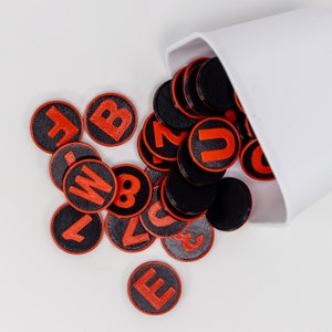 May include: A white cup spills out black and red letter tiles with red letters on a white background. The letters include A, B, C, D, E, F, L, M, N, and O.