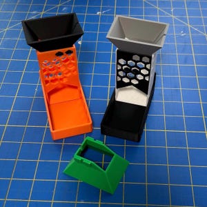 May include: Three 3D-printed plastic objects. Two are vertical, with a funnel top, a honeycomb-patterned midsection, and a tray base. One is orange with a black top, the other is gray with a black base. A green piece sits below.