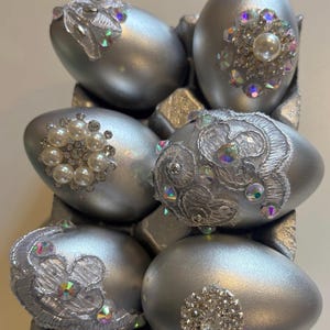 May include: Six silver-painted decorative eggs arranged in a carton. Each egg features unique embellishments including white lace floral appliqués, iridescent rhinestones, and clusters of faux pearls set in silver-toned metal settings.