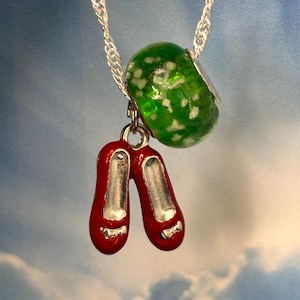 May include: A silver necklace featuring a green glass bead with white speckles and a pair of red enamel shoes with silver details. The shoes have small bows. The necklace is suspended against a sky-blue background.