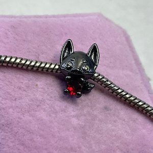 May include: Black charm with a fox design holding a red heart. The charm is made of metal and has a shiny finish.