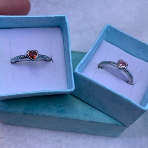 May include: Two silver rings with heart-shaped stones. The ring on the left has a red stone, and the ring on the right has a pink stone. Both rings are in small blue boxes.