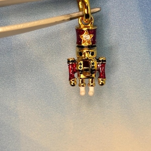 May include: A gold-colored charm in the shape of a toy soldier, held by tweezers. The charm features a red and gold design with a star-shaped accent and a clear gem. The toy soldier has white feet and red arms.