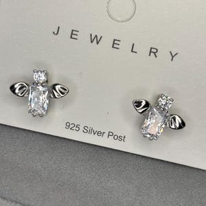 May include: A pair of silver bee earrings with clear cubic zirconia stones. The earrings are on a white card with the word "JEWELRY" printed on it. The card also says "925 Silver Post".