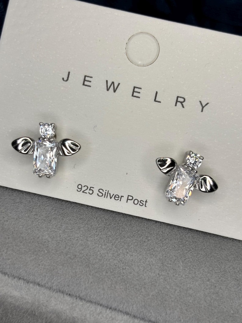 May include: A pair of silver bee earrings with clear cubic zirconia stones. The earrings are on a white card with the word "JEWELRY" printed on it. The card also says "925 Silver Post".