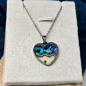 May include: A silver-toned necklace with a heart-shaped pendant. The pendant features a vibrant, iridescent abalone shell design with a mountain scene. The bottom half of the heart is a cream color, and the necklace is displayed on a neutral-toned stand.