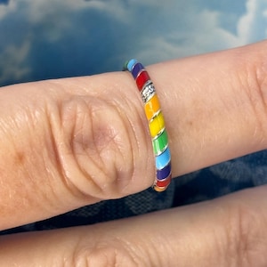 May include: A rainbow-colored ring with a silver band. The ring features a series of vibrant colors: red, orange, yellow, green, blue, and purple. A small, clear stone is set into the ring. The ring is worn on a finger.