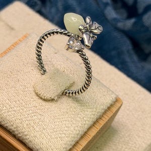 May include: A silver-toned ring featuring a twisted band design. The top of the ring is adorned with a small, pale green, smooth, teardrop-shaped stone and a clear, faceted, triangular-cut stone, both set next to a small silver butterfly motif.