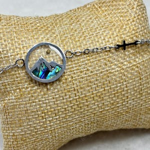 May include: A silver-toned bracelet featuring a circular pendant with a mountain scene and iridescent abalone shell inlay. The bracelet includes a small cross charm and is displayed on a textured, tan surface.