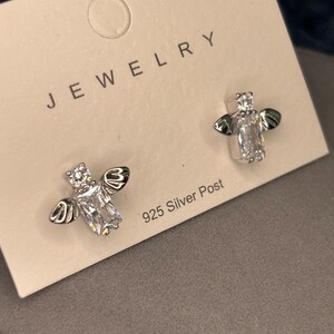 May include: A pair of silver bee earrings with clear cubic zirconia stones. The earrings are on a white card with the word "JEWELRY" printed on it and the text "925 Silver Post".