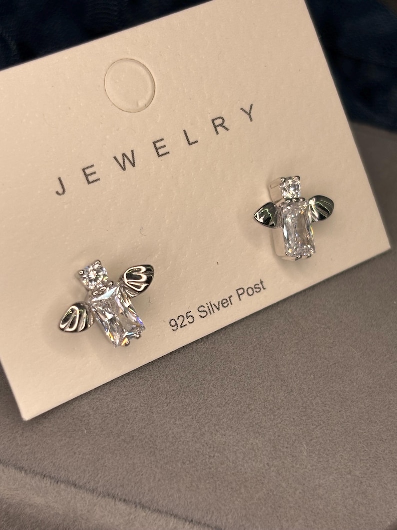 May include: A pair of silver bee earrings with clear cubic zirconia stones. The earrings are on a white card with the word "JEWELRY" printed on it and the text "925 Silver Post".