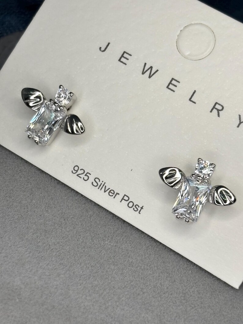 May include: A pair of silver bee earrings with clear cubic zirconia stones. The earrings are on a white card with the word "JEWELRY" and "925 Silver Post" printed on it.