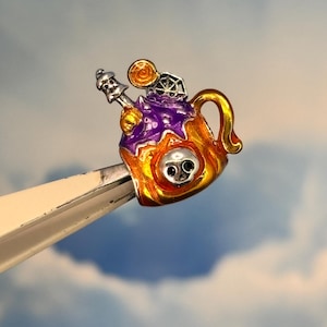 May include: A Halloween-themed charm in the shape of a mug, featuring a skull, spiderweb, and a lollipop. The mug is orange and gold with purple accents. The charm is held by a pair of tweezers against a blue sky background.