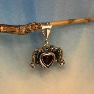 May include: A silver pendant featuring two skeletal hands cupping a central dark red heart-shaped stone. The metal bail is stamped with 925 AG, indicating sterling silver composition. The pendant hangs from a small jump ring attached to a wooden branch.