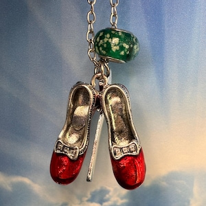 May include: A pendant necklace featuring a pair of red, glittery shoes with silver trim, reminiscent of ruby slippers. The shoes are suspended from a silver chain, with a green and white bead at the top. The background is a blue sky.