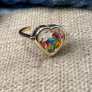 May include: A gold-colored ring with a heart-shaped centerpiece. The heart is filled with colorful, faceted gemstones. The ring is set against a neutral, textured background, with a blue fabric in the background.