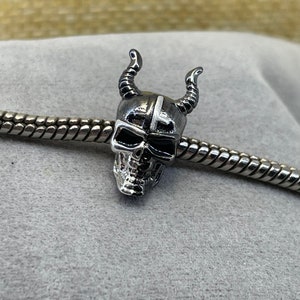 May include: A silver charm with a skull design. The skull is wearing a helmet with horns and a white cross on the front.