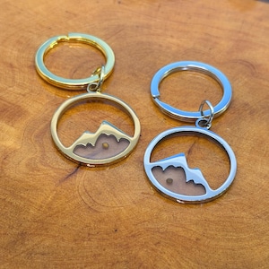 May include: Two keychains with mountain pendants. One keychain is gold-colored, the other is silver-colored. Each pendant features a mountain silhouette within a circular frame, with a small, light-colored object inside. The keychains are on a wooden surface.