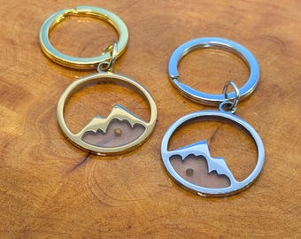 May include: Two keychains with mountain pendants. One keychain is gold-colored, the other is silver-colored. Each pendant features a mountain silhouette within a circular frame, with a small, light-colored object inside. The keychains are on a wooden surface.