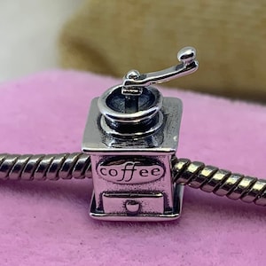May include: Silver charm in the shape of a coffee grinder with a crank handle. The grinder has a drawer and a label that says "coffee".
